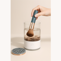 Hand cleaning a makeup brush in an electric brush cleaner with transparent water tank, visible dirt dissolving in water, and detachable silicone lid placed beside the device.