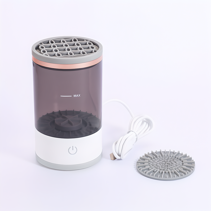 Electric automatic makeup brush cleaner with a detachable transparent rinse tank, gray silicone lid, and included USB charging cable, displayed on a white background with an additional brush cleaning pad.